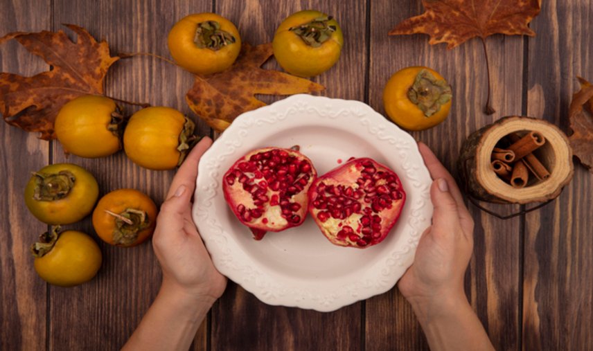 top view of female hands holding a bowl of pomegranates with persimmon fruits isolated on a wooden background
